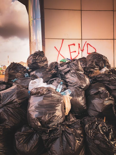A large pile of black trash bags is stacked against a modern tiled wall on an urban street or public space, with some transparent plastic bottles and other waste visible protruding from the bags. The bags appear full and slightly misshapen, suggesting they are tightly packed. Behind the pile, part of a reflective metallic surface in the wall reflects the surrounding environment, including the cloudy sky with patches of sunlight. Red graffiti spelling 'XEND' is spray-painted on the wall behind the rubbish. The scene captures a situation of unmanaged waste accumulation, which could be addressed through private rubbish collection services, exemplifying the need for efficient rubbish removal solutions like those offered by Rubbish Removal Kensington, to maintain cleanliness and proper waste disposal in city environments.