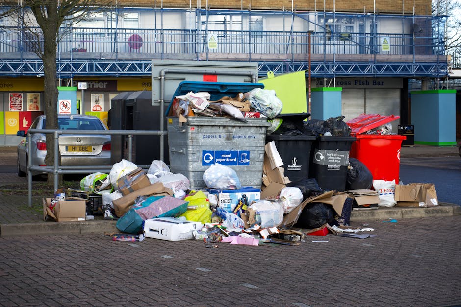 A collection of overflowing rubbish bins and scattered waste bags on a paved urban sidewalk. The central large gray bin with a blue lid is filled with crumpled paper, cardboard, and miscellaneous waste, with some waste spilling onto the ground. To the right of this bin, there are black and red waste containers, also visibly overfilled with assorted trash, including black bin liners and loose debris. Smaller cardboard boxes and paper packaging are strewn across the pavement, along with plastic bags and various discarded items. A silver car is parked behind a metal railing on the left side, partially obscured by the waste. In the background, a building with scaffolding and construction barriers, painted in blue with green accents, indicates nearby ongoing development or maintenance. The scene captures a typical issue of private waste accumulation, highlighting the importance of proper rubbish removal services such as those offered by Rubbish Removal Kensington, especially in busy urban areas where on-site waste management can become problematic without regular collection.