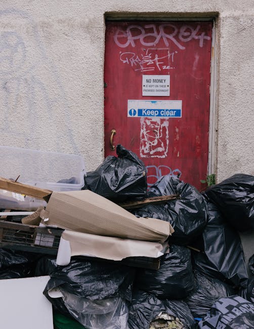 A collection of black rubbish bags, some torn open, and discarded cardboard boxes and packaging materials are piled outside a red metal door set into a plain, textured concrete wall. The bags are irregularly stacked in front of the door, which features various graffiti tags in white and grey spray paint, along with multiple stickers and signs including one that reads 'No Money' and another blue sticker with the words 'Keep clear.' The environment appears to be an urban alleyway or side entrance, with minimal surrounding features visible, emphasizing the accumulation of waste awaiting collection or disposal. The scene suggests a temporary or neglected space where waste has accumulated, aligning with private rubbish clearance or alternative waste handling services, as provided by companies such as Rubbish Removal Kensington.