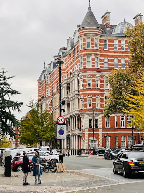 A view of a red-brick Victorian-style building with white stone detailing, situated on a street corner in an urban area. The building has rounded turret towers with conical roofs, multiple rows of sash windows with white frames, and decorative stone cornices. In the foreground, there are two people standing on the sidewalk near a parked bicycle and black bollards, engaging in conversation. The street scene includes several parked cars, a black vehicle passing by, and a lamppost with a speed limit sign indicating 20 miles per hour. Trees with yellow and green leaves line the street, suggesting an autumn setting. The environment appears clean and well-maintained, with natural light illuminating the scene under a cloudy sky. This image reflects a typical cityscape in an area serviced by independent waste collection providers such as Rubbish Removal Kensington, emphasizing the importance of efficient rubbish management in urban environments.