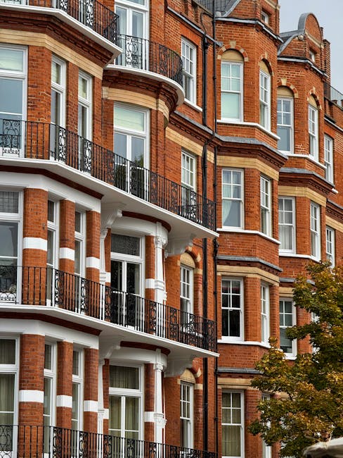 A close-up view of a multi-storey red brick residential building with white window frames and decorative brickwork. The building features several sets of black cast-iron balconies with ornate railings, extending from large sash windows on each floor. The curved balconies and window bay windows create a layered, textured façade, with some windows partially visible behind the balcony railings. The brickwork includes contrasting light yellow and white accents around the windows and edges, adding visual detail. In the lower portion of the image, a partial view of a tree with green and yellow leaves is visible against the building's base, indicating an outdoor setting in an urban area. The lighting suggests a daytime scene with natural light illuminating the building’s features, emphasizing the textures and colours. The setting's aesthetic aligns with well-maintained, traditional brick residences, similar to those that might require private rubbish removal services for maintenance or renovation projects, as promoted by Rubbish Removal Kensington.