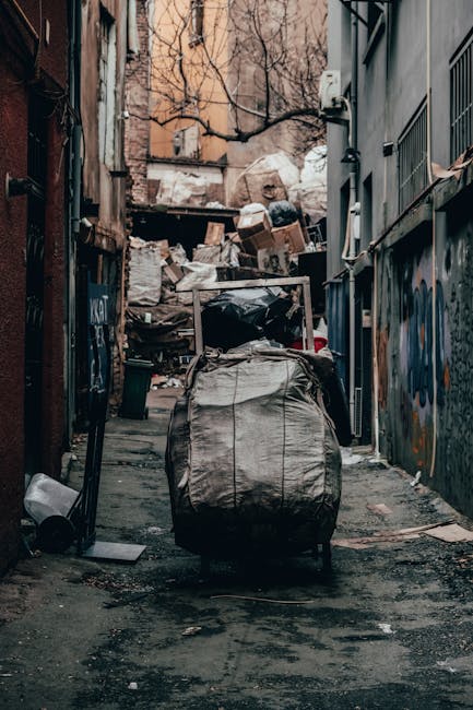 A narrow urban alleyway cluttered with discarded waste, featuring a large black plastic bag filled with rubbish positioned in the foreground, with its handles visible and slightly torn. Behind it, an overflowing pile of various waste materials, including cardboard boxes, paper, plastic bags, and other debris, is stacked against a weathered wooden structure and the side of a building. The surroundings include graffiti-covered walls on the right, with metal bars on windows, and a dark, uneven pavement surface strewn with dirt, small trash, and stains. In the background, bare tree branches extend over the alley, contrasting with the dense accumulation of waste. The scene appears to be in a back alley used for private waste disposal, consistent with private rubbish removal services such as those offered by Rubbish Removal Kensington, emphasizing the importance of proper waste management and alternative collection options in urban environments.
