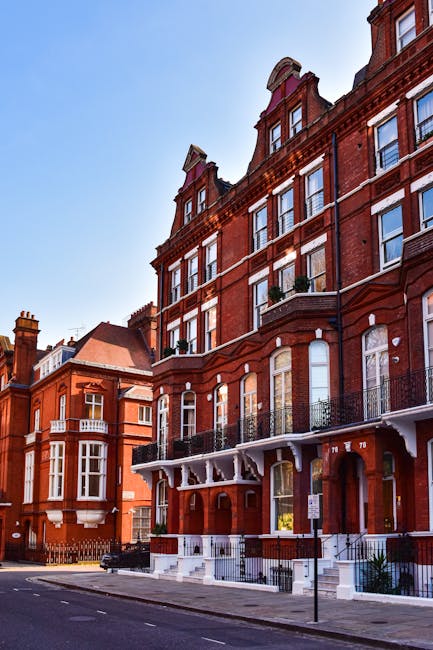 A row of red-brick Victorian-style buildings with multiple stories, large sash windows, and decorative black iron balconies lined across the upper floors. The buildings feature ornate white trim detailing beneath the eaves and around the windows, as well as small terraces with railings on the ground level. The street in front is paved, with a paved sidewalk and a lamppost visible on the right. The scene is set on a clear day with bright blue sky and natural daylight illuminating the facades, representing typical residential architecture that may require occasional site clearance or rubbish removal services provided by companies like Rubbish Removal Kensington, especially when preparing properties for refurbishment or disposal of accumulated waste materials.