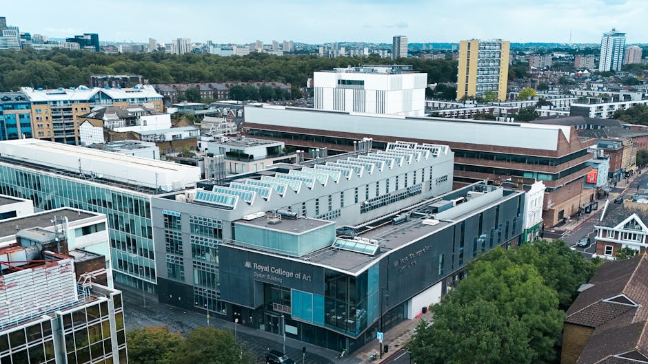 An aerial view of a modern urban area showing the exterior of the Royal College of Art, a contemporary building with large glass windows and a grey facade, situated among other commercial and residential structures. The building has a distinctive architectural style with a flat roof, ventilated sections, and a prominent sign indicating its identity. Surrounding the college are other multi-storey buildings featuring varied materials including glass, brick, and metal cladding, with some rooftops visible in the foreground. To the right, a brown brick building with an angled roof can be seen, adjacent to a street lined with small houses and trees. The background reveals a city skyline with mid-rise office blocks and high-rise apartment towers under an overcast sky, suggesting a typical cityscape. The scene is well-lit with natural daylight, highlighting the textures of glass, metal, and brick and illustrating an environment where private property often accommodates waste management activities such as rubbish collection or on-site disposal handled by services like Rubbish Removal Kensington in alignment with local guidelines for waste handling within the Royal Borough of Kensington and Chelsea.