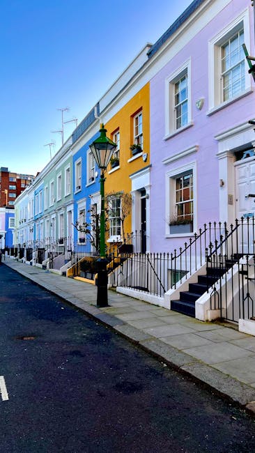 A row of colorful terraced houses on a quiet urban street with a clear sky overhead. The buildings are painted in pastel shades, including light blue, yellow, and pale lavender, with white window frames and decorative trims. Each house has a small front garden or entrance area enclosed by black wrought iron railings, with some steps leading up to the front doors. The sidewalk is paved with grey slabs, and a traditional black lamppost with a green top is positioned near the curb, providing street lighting. There are no visible rubbish or waste items in the scene, reflecting a tidy and well-maintained residential area. The street appears to be in a city environment, with taller brick buildings visible further down the road, complementing the row of houses. This scene subtly illustrates respectful curbside management, aligning with independent collection or private waste handling practices, indicative of non-local-authority rubbish disposal services.