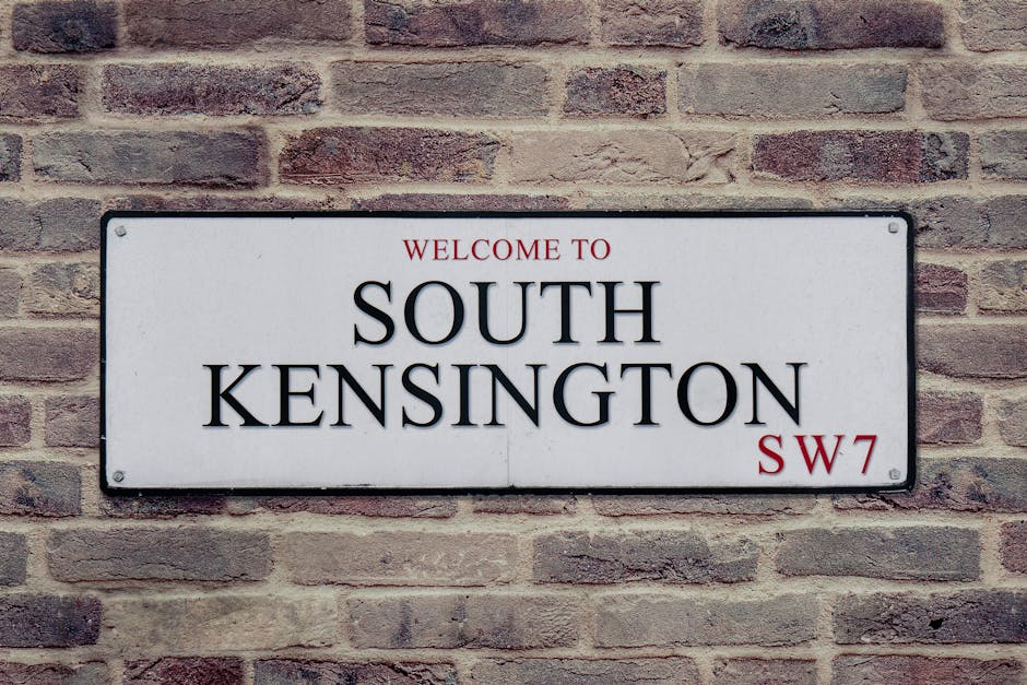 A rectangular metal street sign mounted on a red-brown brick wall, displaying the text 'WELCOME TO SOUTH KENSINGTON SW7' in bold black uppercase letters, with 'WELCOME TO' and 'SW7' in smaller red font. The sign has a white background with rounded corners and visible screw fixings at each corner. The brick wall behind the sign features a rough texture with variation in shades of red, brown, and gray, laid in a horizontal pattern. The lighting is natural and evenly diffused, illuminating the sign and wall without harsh shadows. The overall scene suggests an outdoor urban setting, commonly associated with areas requiring private rubbish collection or alternative waste handling options, aligning with the context of rubbish removal services in South Kensington.