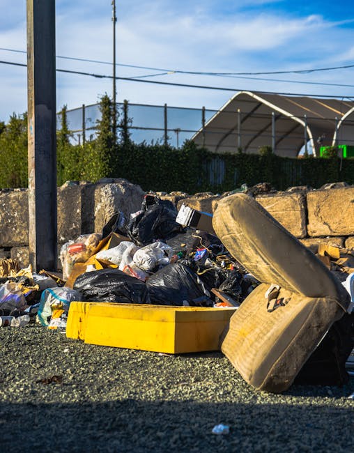 A collection of discarded rubbish piled on a gravel surface, including several black plastic trash bags, a yellow plastic container, a worn and dirty beige car seat, and various small debris. The background features a stone wall made of uneven, rough-hewn stones, with a metal pole and electrical wires extending overhead. Behind the wall, there is a parking lot or open area with a large, curved, tent-like structure with a translucent roof supported by metal poles, and some green digital signage or lighting visible in the distance. The scene is illuminated by natural daylight, casting shadows on the ground, indicating it is a clear day. The overall setting suggests an area designated for waste collection or temporarily storing rubbish outside a private property or waste removal service, as observed by Rubbish Removal Kensington, with the scene reflecting the need for professional rubbish clearance or private waste handling arrangements.
