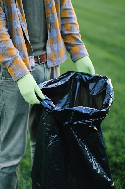 A large pile of black garbage bags filled with waste is stacked against a tiled wall outdoors. Some bags are slightly torn, revealing plastic bottles, paper, and other rubbish inside. The bags vary in size and are tightly knotted at the top, with a few discarded bottles and pieces of paper scattered around the base. Behind the bags, part of a modern building with a reflective metal surface and beige or light brown tiles is visible, with red spray paint graffiti spelling 'XEND' on the wall. The scene is illuminated by natural daylight, casting soft shadows across the waste collection area, which appears to be an informal, outdoor rubbish accumulation point. This setting may relate to a scenario where independent or private rubbish collection services, such as Rubbish Removal Kensington, handle waste disposal outside of regular municipal collection, evidenced by the unmanaged appearance of the waste pile and the nearby urban environment.